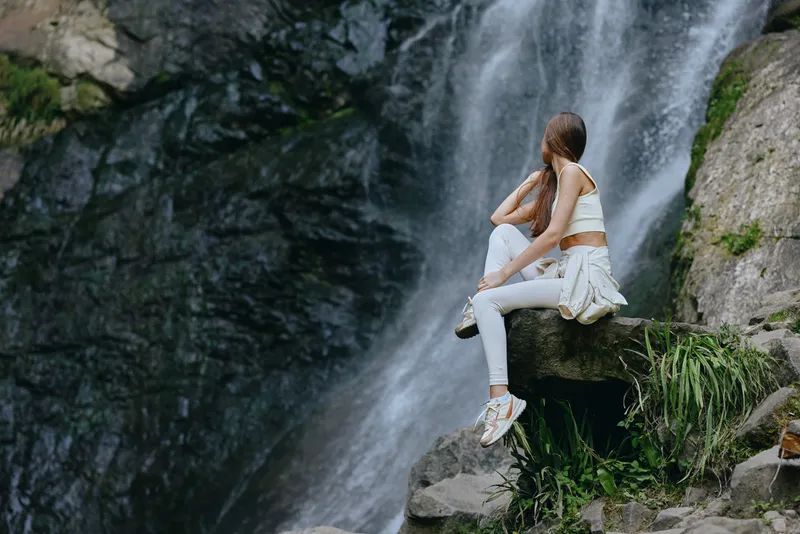 Young woman sitting on rock near waterfall, wearing white casual clothes and sneakers, enjoying nature and peaceful outdoor environment during daytime.