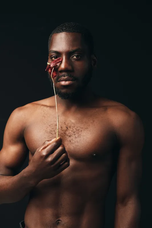 Black man with dark skin and muscular body holding dried red flower near face in creative visual designer style on black background. Artistic portrait with serious expression and bare chest.