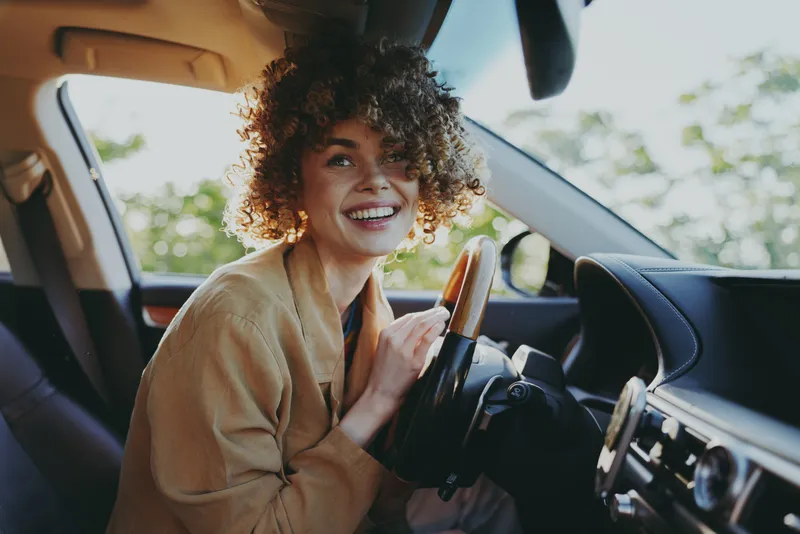 Woman lifestyle portrait inside a car, smiling driver with curly hair, warm retro film color mood, casual outfit, natural light, positive vibe, focused on steering wheel and dashboard