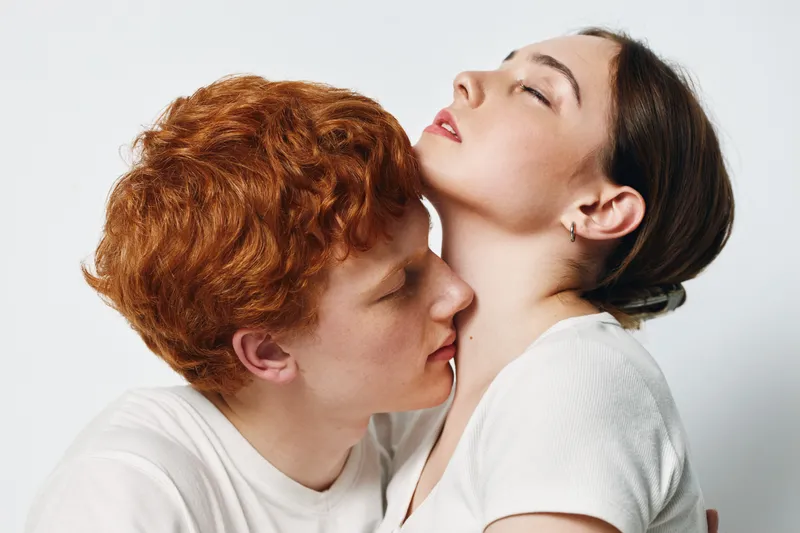 Couple, affection and intimacy with young man kissing woman neck in studio. Redhead male and brunette female in white clothes against plain background with love moment.