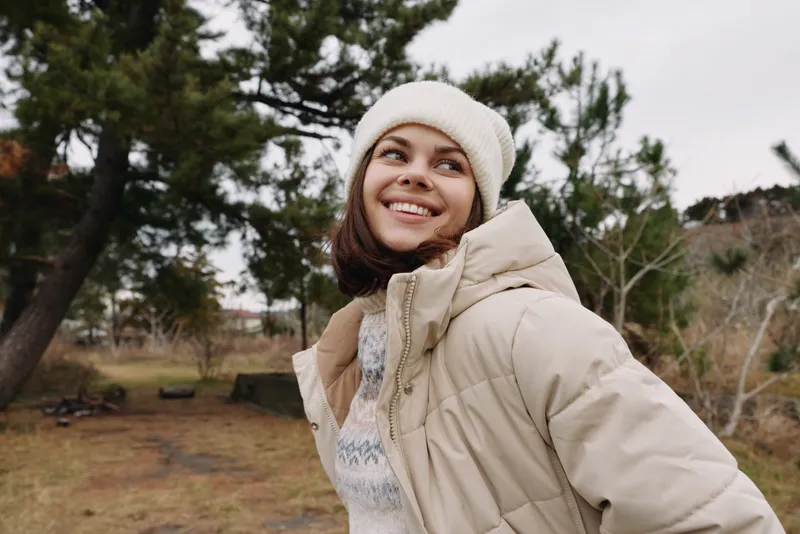 Woman posing outdoors in winter wearing a puffy jacket and knit beanie, smiling as bare trees and a park hillside create a calm, scenic background for a lifestyle portrait