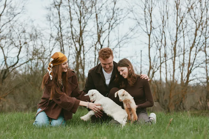 Three friends outdoors in a grassy field with two goat kids, smiling and petting them. Casual clothing, warm mood, nature setting, playful interaction.