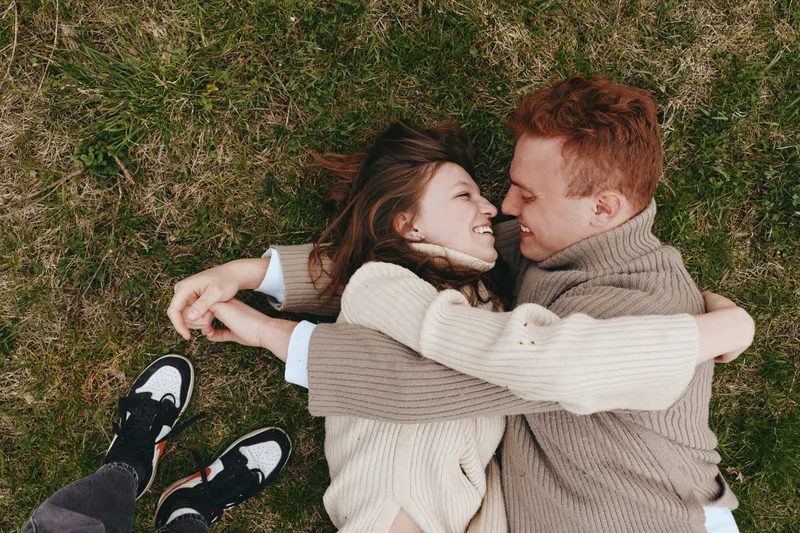 Couple outdoors lying embrace, smiling and relaxed, sunlit moment on grass, intimate connection, warm clothing, casual style, candid mood, natural colors, lush greenery background, portraying comfort