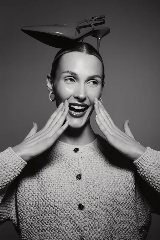 Young caucasian woman with fair skin and dark hair laughs with hands near face in studio. Creative concept with high heel shoe on head and expressive joyful mood.
