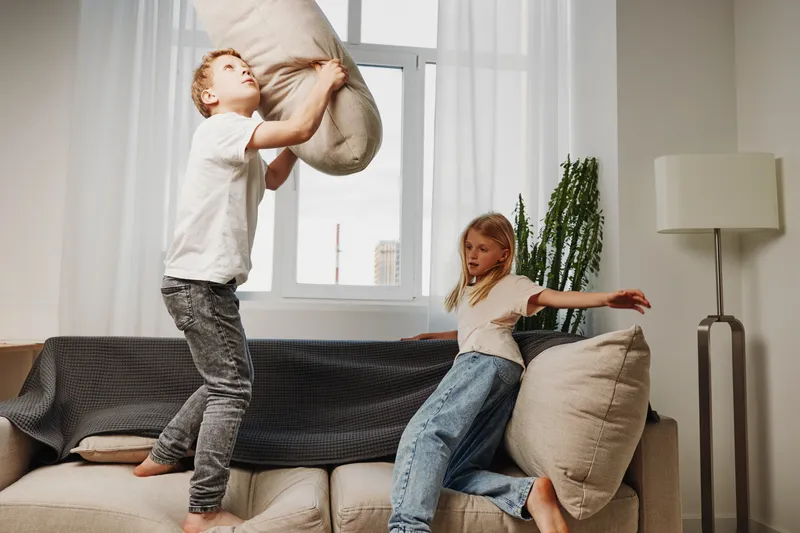 Two children having fun pillow fighting on a sofa in a bright living room with natural light, capturing joyful family moments and playful childhood energy indoors.