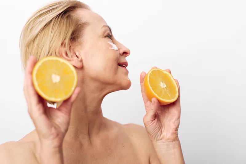 Mature woman with natural skin holds two orange halves near face smiling with cream on cheek isolated on white background. Concept of skincare, healthy lifestyle, and natural beauty.