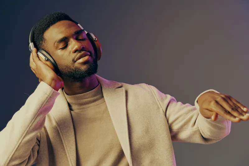 Young Black man enjoying music with headphones, wearing a stylish beige sweater, against a gradient purple background, conveying relaxation and joy