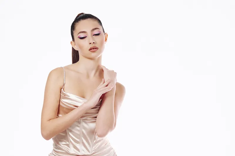 Elegant young woman wearing a satin dress poses with eyes closed and hand touching her arm. High fashion makeup and sleek hairstyle against a white background. Concept of beauty and creativity.