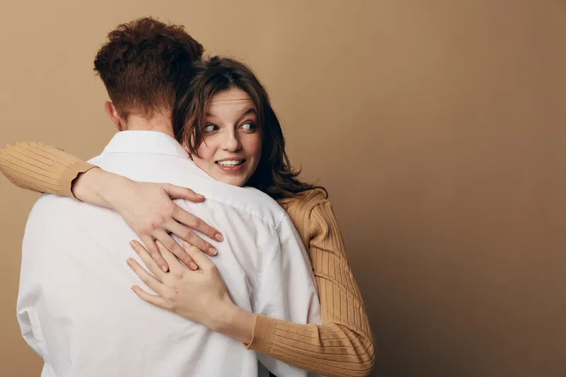 Young woman in brown sweater hugs man in white shirt, looking surprised and happy. Intimate moment with warm beige background, concept of affection, relationship, and emotional connection.