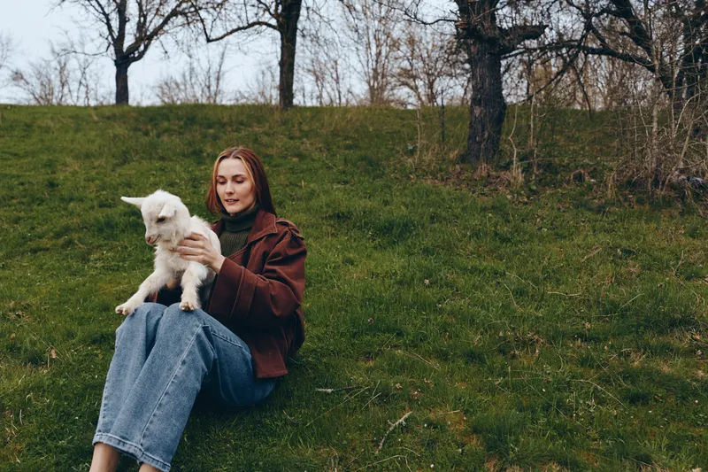 Young woman with red hair sits on a grassy hill holding a white baby goat, wearing a brown jacket and blue jeans in a calm outdoor lifestyle setting.