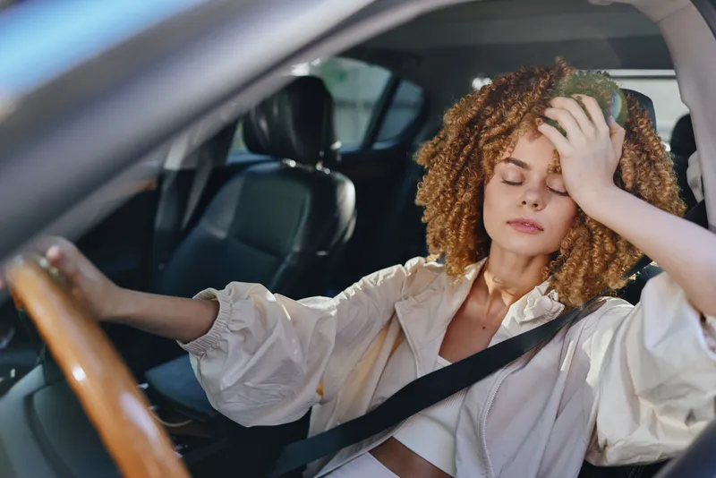tired woman sitting in car holding ice pack on forehead, experiencing fatigue and heat, young curly-haired woman wearing light blouse feeling unwell.