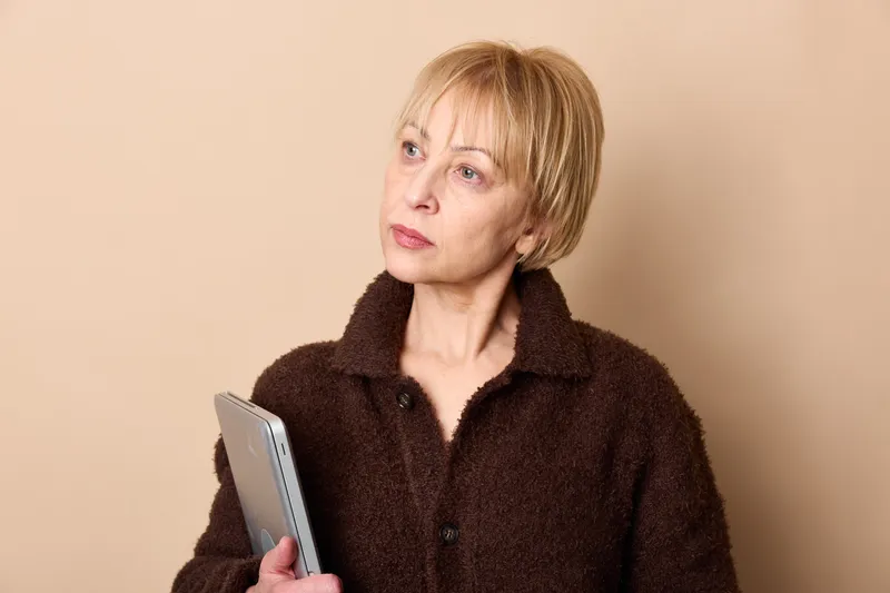 Woman with smartphone and communication, mature blond hair, studio portrait on beige background, looking away thoughtfully, calm expression, indoor lighting, closeup