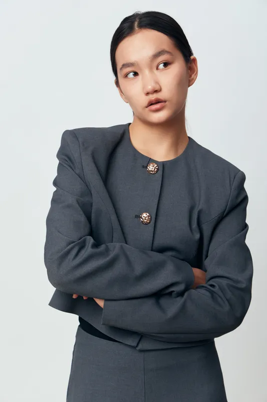 Young Asian woman in gray formal suit standing with crossed arms looking sideways, portrait of confident business professional in studio with neutral background and minimalistic style.