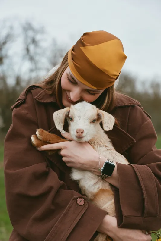 Goat, kid, woman, outdoors, animal, cuddle, lifestyle, countryside, a warm moment as a smiling woman holds a young goat against her brown coat in a peaceful rural setting, expressing tenderness and
