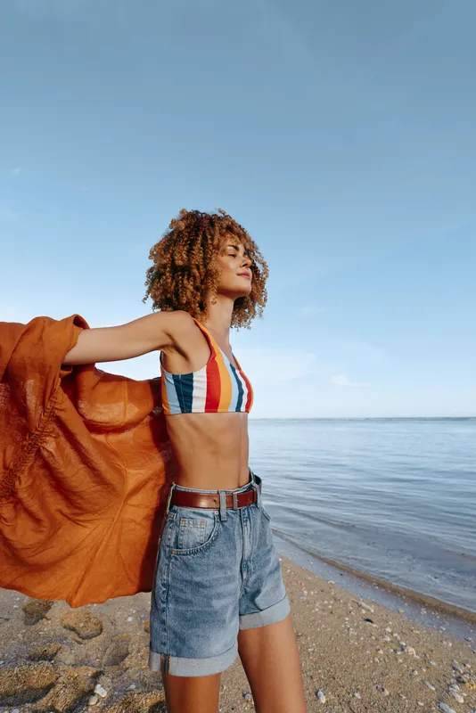 A youthful woman on a sunny beach, striped bikini top and denim shorts, holding an orange towel.