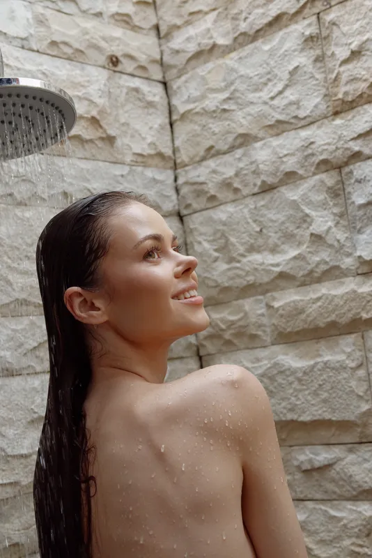 young woman enjoying shower with wet hair under indoor showerhead, stone wall background, natural skincare and relaxation concept