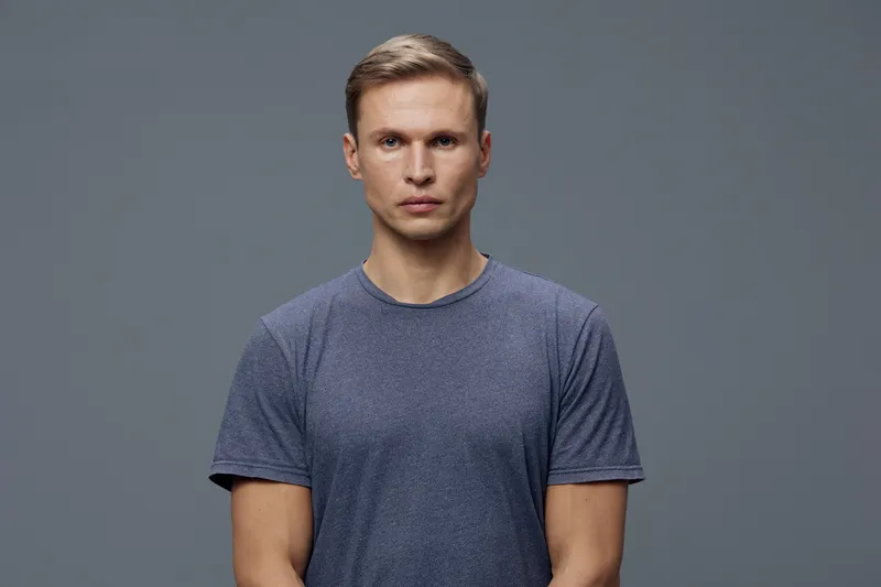 Portrait of a young man with short blond hair and serious expression, wearing a casual blue t-shirt, looking directly at camera, studio shot with neutral gray background, minimalistic style. People li