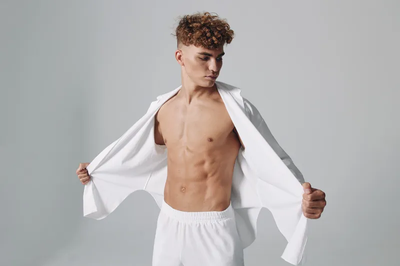 Young man with curly hair posing shirtless in white pants and open white shirt against gray background. Muscular torso, confident expression and casual style in studio lighting.