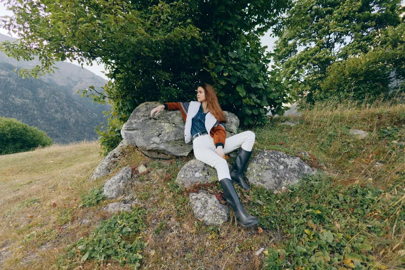 Woman in nature on rock under tree beside mountains, wearing boots and stylish fashion layers, reclining and looking away in outdoor pastoral scene with lush green landscape.