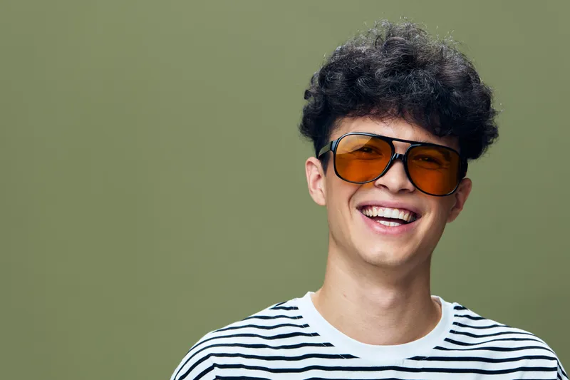young man wearing orange sunglasses smiling brightly with curly hair and striped shirt against green background, casual style portrait with cheerful mood