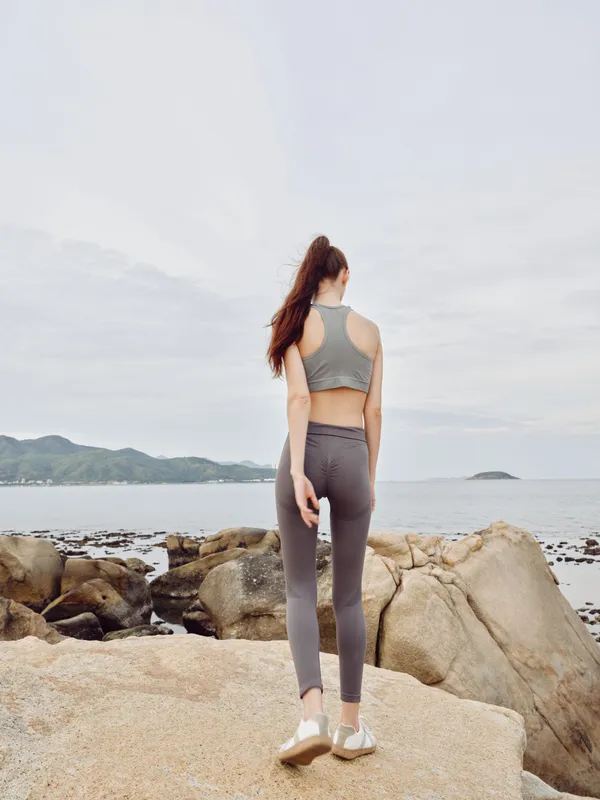 Young woman in activewear enjoying nature by the ocean, reflecting a healthy lifestyle and fitness concept with a serene backdrop of rocks and water