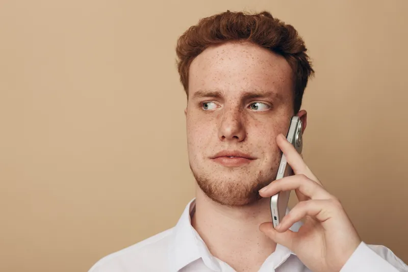 Young man with red hair and freckles wearing white shirt talks on smartphone looking sideways. Neutral beige background. Concept of communication, technology, and casual lifestyle.