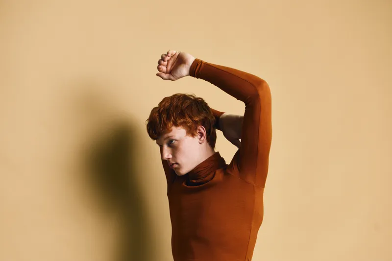 Young man with red hair wearing brown turtleneck sweater stretching arms against beige background with strong shadow on wall in studio.