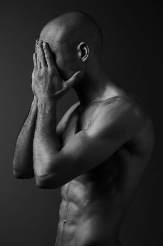 Black and white image of a contemplative man covering his face, showcasing emotions of introspection and vulnerability, set against a dark background.