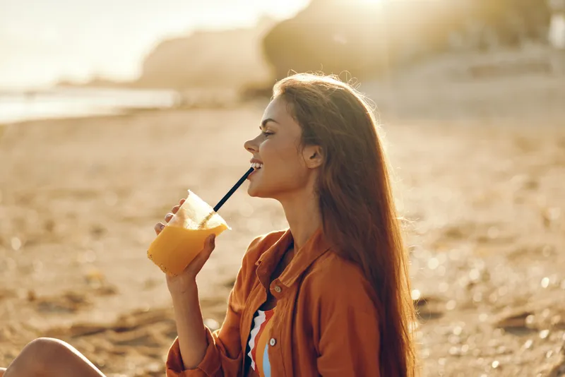 Woman outdoors drinking straw beverage in warm sunlight, smiling in side profile, enjoying golden hour, light breeze, relaxed posture, sandy beach background, soft natural colors today