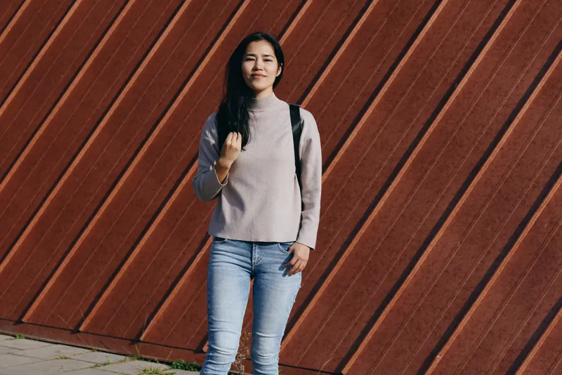 Woman standing in a casual sweater and jeans beside a diagonal wooden wall outdoors, backpack over shoulder, relaxed pose, daylight fashion mood, simple contemporary composition.