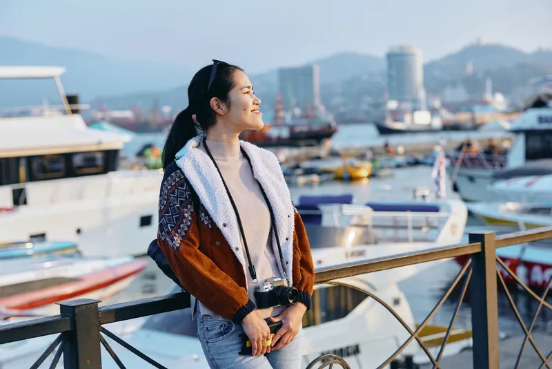 Woman standing by a harbor with boats in the background, wearing a stylish fleece and warm coat, enjoying a sunny day by the water, leisure travel mood, candid moment, seaside scenery and urban