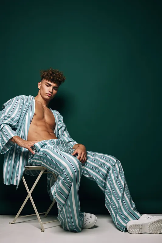 Young man with curly hair wearing striped pajamas and white sneakers sitting on a chair against dark green background in studio. Casual fashion and relaxed pose.