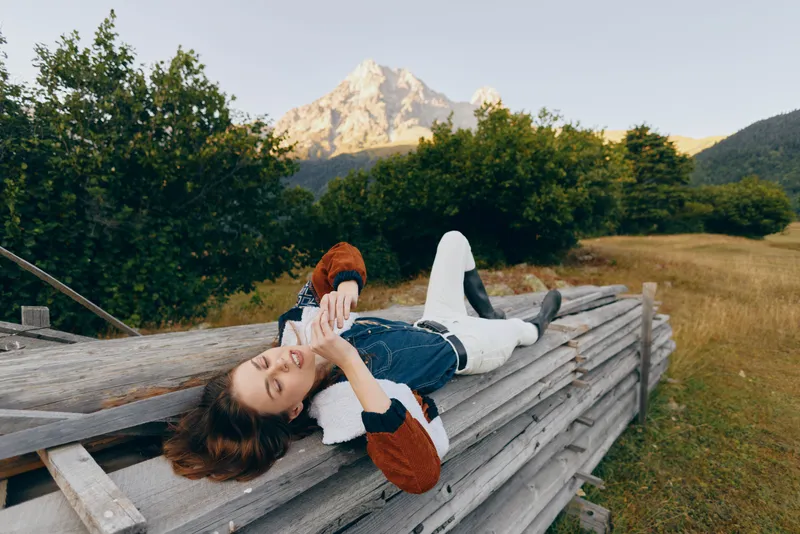 Woman on wooden fence using phone, relaxing in meadow with mountains in background and countryside nature. Casual sweater, rural leisure and peaceful outdoor lifestyle portrait.