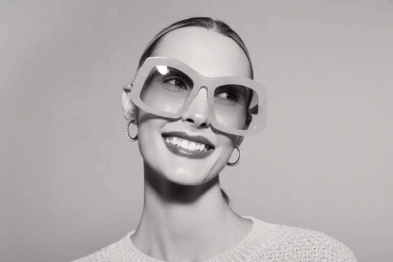 Smiling white woman with fair skin and slicked back hair wearing oversized glasses and hoop earrings. Studio portrait with soft lighting on neutral background. Concept for fashion and eyewear.