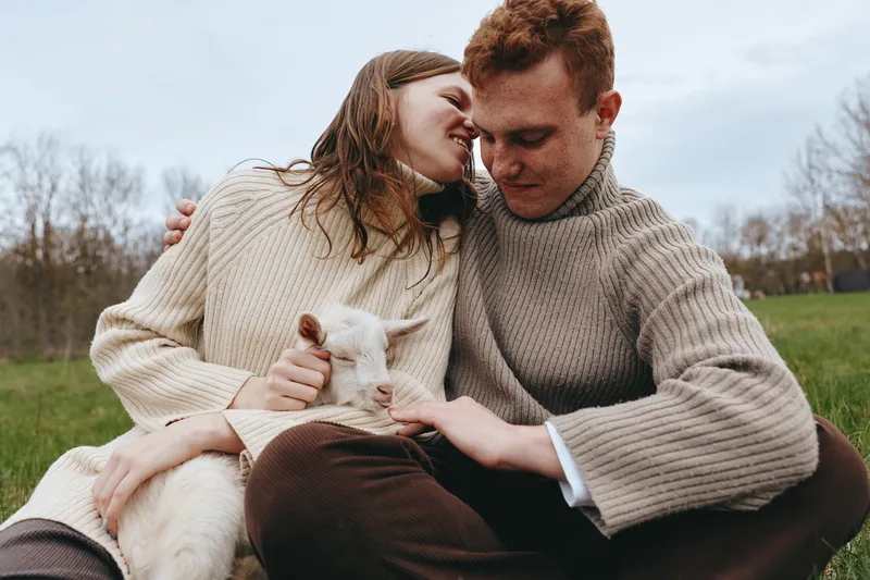Young couple in cozy sweaters cuddle a small white baby goat while sitting on grass in a peaceful outdoor setting during a cool day.