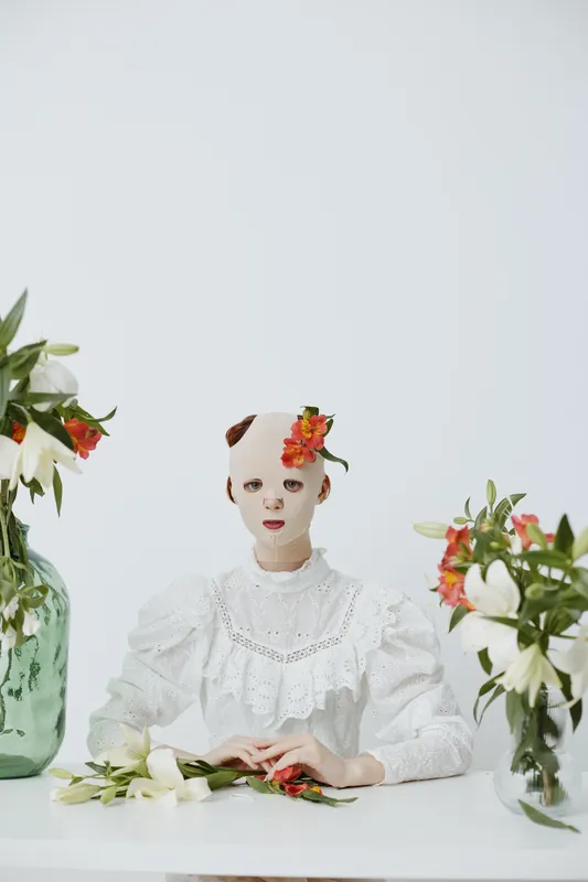 A surreal portrait of a model in a white dress with a mask, surrounded by flowers, conveying a mysterious emotional tone Perfect for conceptual art and fashion