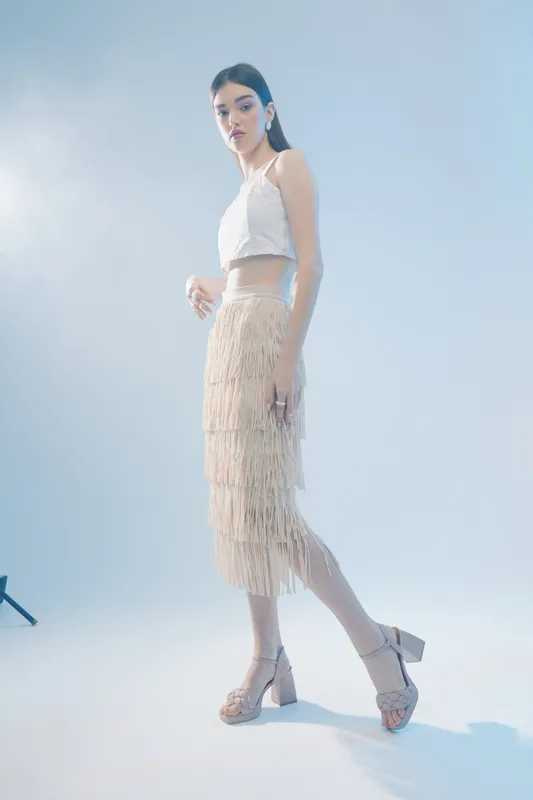 Tall woman with long hair wearing a white top and beige fringe skirt posing in a studio with soft lighting. Concept of woman beauty fashion and elegance.