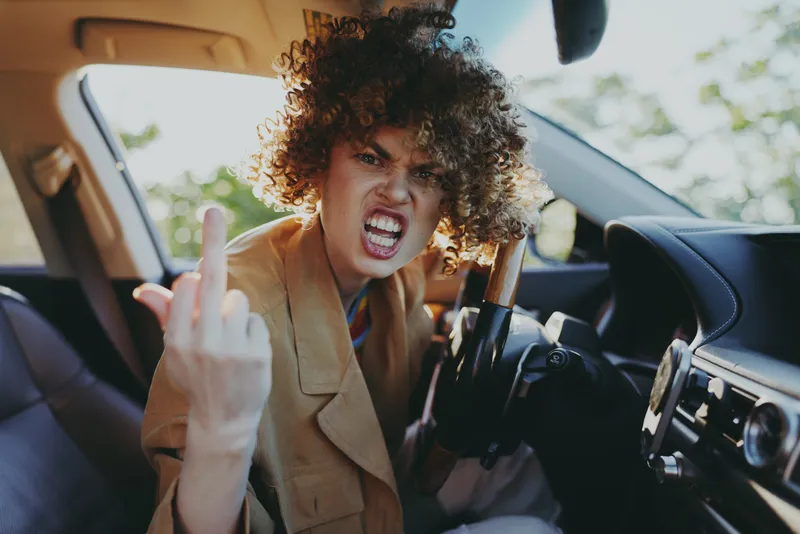 A young woman with curly hair sits in a sunlit car, angrily flipping the middle finger toward the camera, conveying bold rebellion in a candid retro vibe.