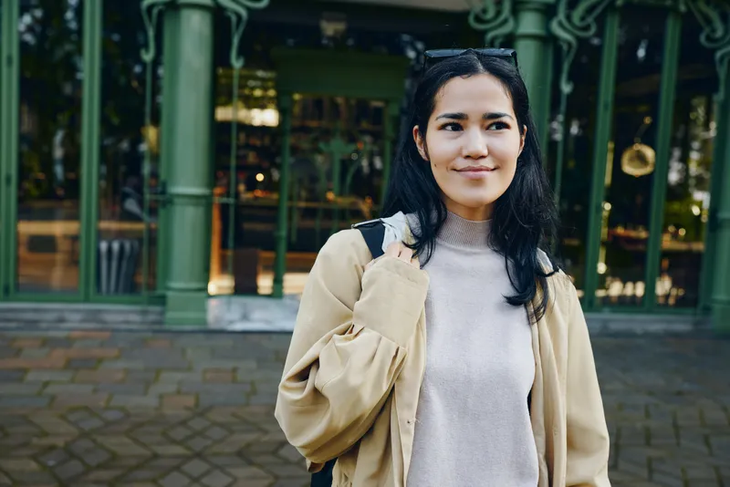 Woman outdoors in casual coat and sweater with a backpack stands on a street, smiling, daylight glow, urban vibe, candid fashion moment, approachable and relaxed