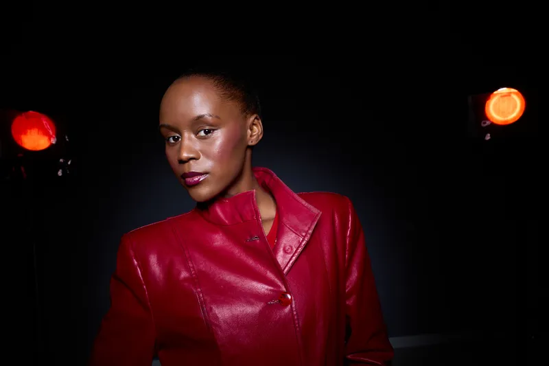 Confident dark skinned African woman wearing a red leather jacket poses against dark background with bright orange lights. Concept of fashion, style, model, and portrait photography.