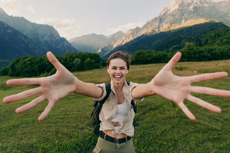 Happy woman outdoors reaching hands forward smiling with mountains and green meadow in background casual outfit natural light joyful lifestyle moment