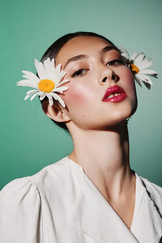 Young woman with bold red lips and glowing skin wears daisies as earrings against pastel background. Fashion beauty portrait with creative floral styling and fresh look.