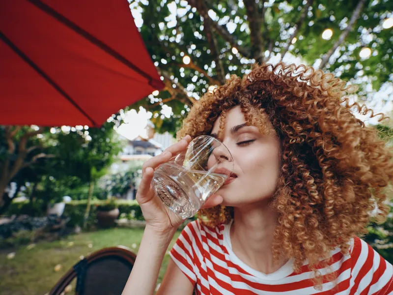 A woman with curly hair drinks water from a glass outdoors under a red umbrella, in a sunlit garden cafe, capturing a refreshing, casual daytime moment.