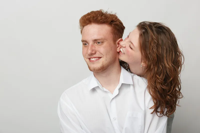 Happy young couple in white shirts share affectionate moment, woman whispering in man’s ear. Studio portrait on light background, concept of love, intimacy, and relationship happiness.