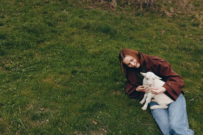 Young woman in a brown jacket sits on green grass holding a white baby goat in her arms in a peaceful outdoor lifestyle scene.