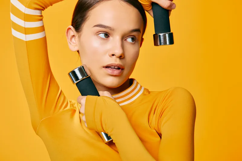 Fitness woman in a bright yellow outfit holds dumbbells against a vivid orange backdrop, showcasing strength, training and a bold sport fashion lifestyle.