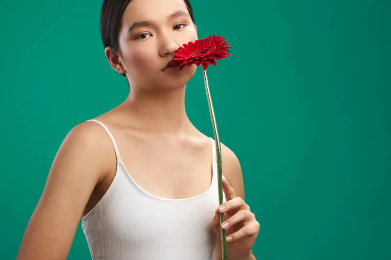 Asian woman holding a red flower near her lips against a green studio backdrop, presenting a calm, intimate moment with soft lighting and a minimalist mood.