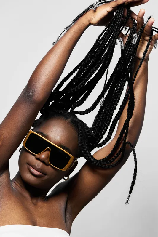Confident black woman with braids holding her hair high, sporting stylish sunglasses, highlighting beauty and self expression in a minimalist setting