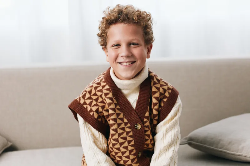 Smiling young boy sits on a sofa wearing a knitted geometric vest over a cozy turtleneck, soft natural light framing a warm, inviting home moment.