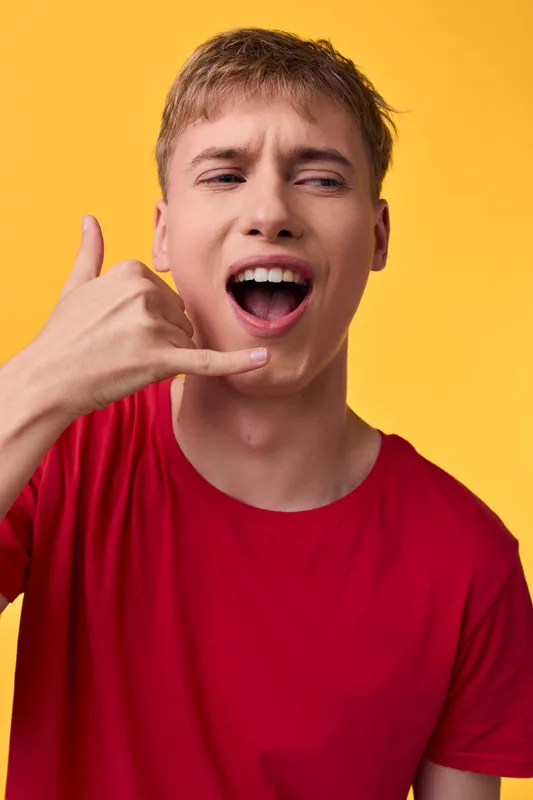 Man in red shirt makes a call gesture against a bright yellow background, conveying communication, urgency, and friendly energy suitable for messaging, customer engagement, and casual business